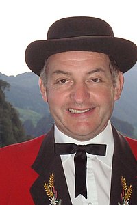 Ein Mann in traditioneller Tracht mit einem schwarzen Hut und rotem Jackett lächelt vor einer bergigen Landschaft.