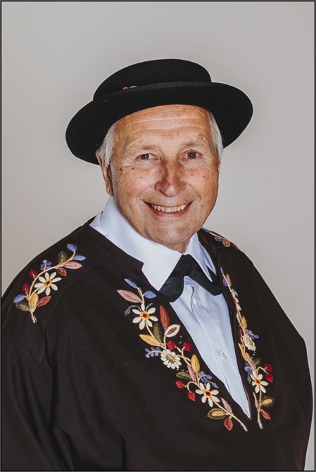 Ein älterer Mann mit einem schwarzen Hut und einem festlichen, bestickten Oberteil lächelt in die Kamera.
