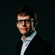 Ein Mann mit Brille und Anzug steht vor einem dunklen Hintergrund.