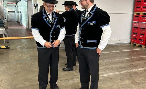 Zwei Männer in traditionellen schwarzen Anzügen mit blauen Akzenten und Hüten stehen in einer Lagerhalle.