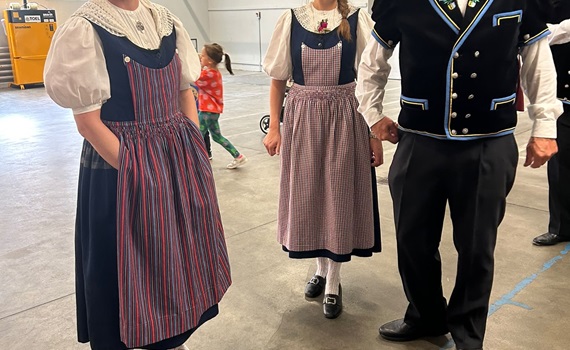 Drei Personen in traditioneller Kleidung stehen in einer Halle, während ein Kind im Hintergrund spielt.