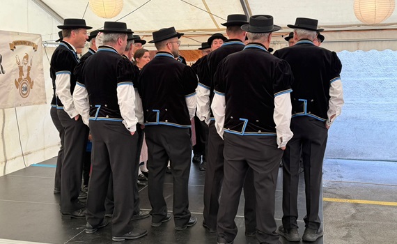 Eine Gruppe von Männern in traditionellen schwarzen Anzügen mit Hüten steht auf einer Bühne in einem Zelt.