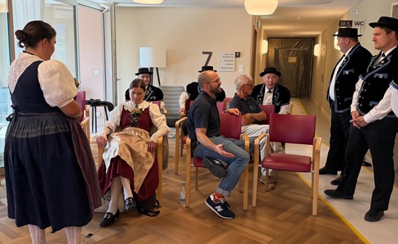Eine Gruppe von Menschen in traditioneller Kleidung sitzt und steht in einem Raum mit Holzfußboden, während eine Person mit einem Mikrofon spricht.