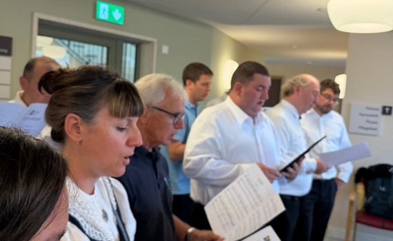 Eine Gruppe von Menschen singt und hält Notenblätter in einer hellen Innenraumumgebung.