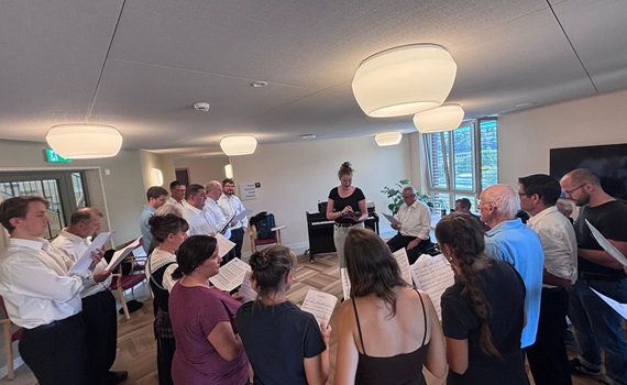 Eine Gruppe von Menschen singt in einem Raum, während eine Person vorne Anweisungen gibt.